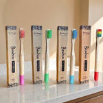 Six bamboo toothbrushes with colorful bristles and matching pink, green, blue, and red handles are lined up next to their kraft paper packaging boxes on a marble countertop.
