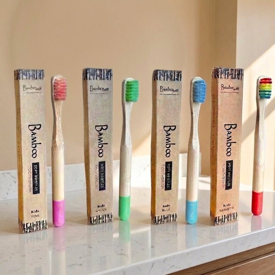 Six bamboo toothbrushes with colorful bristles and matching pink, green, blue, and red handles are lined up next to their kraft paper packaging boxes on a marble countertop.